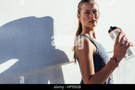 Colpo di bella giovane donna in piedi all'aperto tenendo la bottiglia di acqua e guardando lontano. Femmina Fitness prendendo una pausa dopo allenamento. Foto Stock