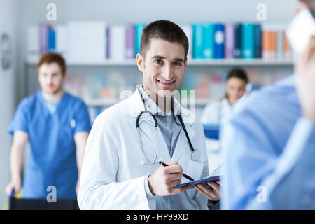 Controllo medico i record medici e tenendo un clipboard, sorride alla fotocamera, personale medico sullo sfondo Foto Stock