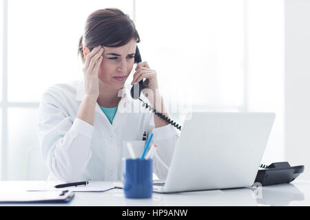 Sottolineato medico donna lavora alla scrivania in ufficio e rispondere a chiamate telefoniche, sta avendo un mal di testa e toccando il suo tempio Foto Stock