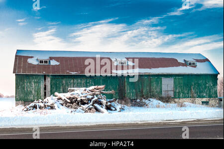 Il vecchio fienile verde sulla fattoria con pila di logs di fronte che sono coperti di neve. lungo l'autostrada in Ontario Canada Foto Stock