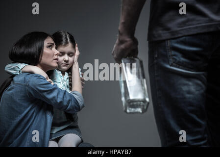 Essere gentili. Spaventata donna abbracciando la figlia di mettere la sua mano sulla testa della sua figlia guardando il suo marito crudele, isolato su grigio Foto Stock