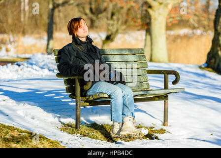 Giovane femmina adulta vestita di nero e blu, crogiolarsi al sole su una panchina nel parco in inverno o in primavera. Neve sulla terra. Soleggiata giornata calda con som Foto Stock