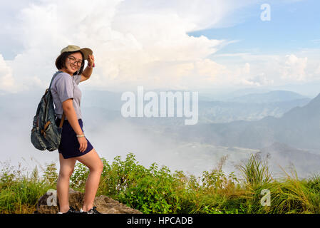 Tourist teens ragazza escursionista con zaino cappello e occhiali è sorriso permanente e pone felicemente in alta montagna su sky e lo sfondo di nebbia al punto panoramico Foto Stock