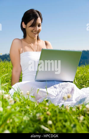 Modello rilasciato , Junge Frau sitzt mit Laptop in der Wiese - donna utilizzando laptop in Prato Foto Stock