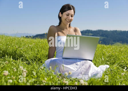 Modello rilasciato , Junge Frau sitzt mit Laptop in der Wiese - donna utilizzando laptop in Prato Foto Stock