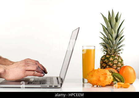 Giovane che lavora su un computer portatile nei pressi di frutta tropicale e un bicchiere di succo di arancia. Foto Stock
