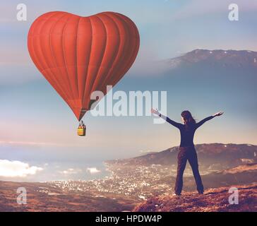 Ragazza con fatto alzare le mani sulla cima della montagna guardando Big Red in pallone aerostatico forma di cuore che vola in cielo blu. Romantica per il giorno di San Valentino. Sport e rec Foto Stock