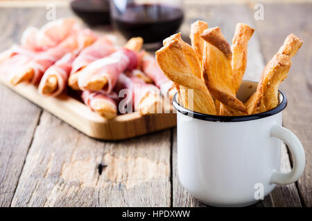 Bastoncini di pane con formaggio di pasta sfoglia in bianco rustico mug sul prosciutto crudo e bicchiere di vino rosso sfondo su un tavolo di legno, deliziosi snack Foto Stock