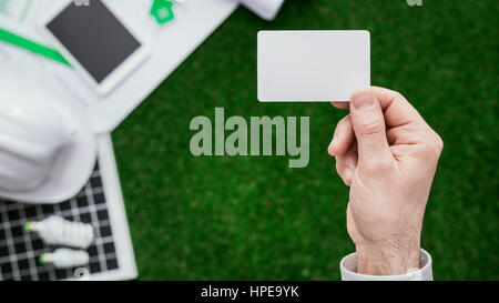 Contraente tenendo un blank business card con progetto di casa, pannello solare ed erba sullo sfondo, verde home il rinnovamento e il concetto di business Foto Stock