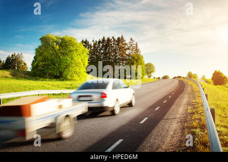 Strada asfaltata sul campo di tarassaco con una vettura con rimorchio. veicolo in movimento sulla serata di sole Foto Stock