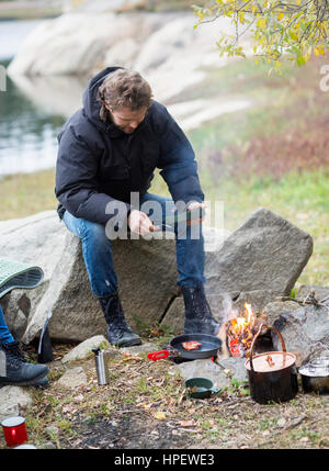 Per tutta la lunghezza della giovane uomo avente il cibo mentre è seduto sulla roccia al campeggio Foto Stock