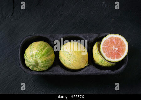 Intero e fette di agrumi rosa limone tigre di carta nella casella di mercato su pietra nera di ardesia di sfondo a trama. Vista da sopra con lo spazio. Moderno mangiare sano Foto Stock