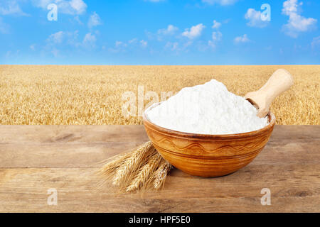 Farina di grano tenero in vaso in ceramica. Spighe di grano e farina di frumento nella ciotola sul tavolo con campo di grano sullo sfondo. Golden campo di grano e cielo blu Foto Stock