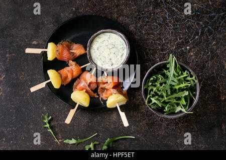 Delizioso antipasto con affumicato con salmone e patate lesse su spiedini servita con cremosa salsa di aneto e rucola sulla piastra nera su backgr scuro Foto Stock