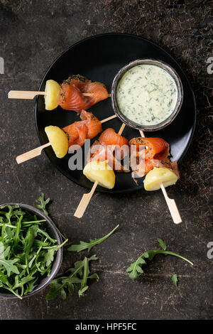 Delizioso antipasto con affumicato con salmone e patate lesse su spiedini servita con cremosa salsa di aneto e rucola sulla piastra nera su backgr scuro Foto Stock