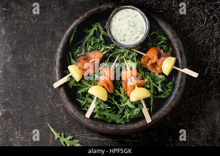 Delizioso antipasto con affumicato con salmone e patate lesse su spiedini servita con cremosa salsa di aneto e rucola in lastra di pietra su backgr scuro Foto Stock