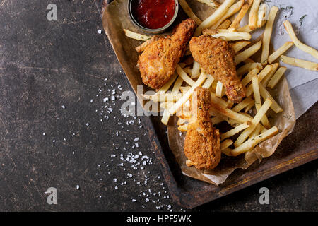 Il fast food fritto croccante di cosce di pollo e patatine fritte di patate con sale e ketchup salsa servita sulla carta da forno nel vecchio forno arrugginito il vassoio su testo scuro Foto Stock