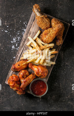 Fast food croccante fritti speziati e le cosce di pollo, le ali e le patatine fritte di patate con sale e ketchup salsa in legno che serve scheda sopra te scuro Foto Stock