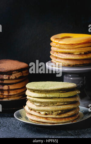 Varietà di fatti in casa american ombre cioccolato, tè verde Matcha e curcuma frittelle con miele e salsa servita su piatti e una fetta di torta in piedi sopra il nero s Foto Stock