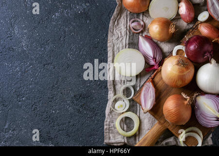 Varietà di interi e fette di rosso, bianco, giallo e scalogno cipolle sul legno taglio chopping board sul tessile igienico su pietra scura backgroun texture Foto Stock