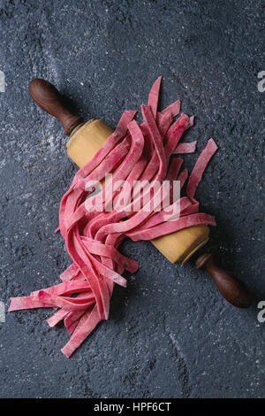 Crudo fresco non cotti in casa rosa barbabietola tagliatelle di pasta di legno sul perno di rotolamento su dark texture di sfondo di calcestruzzo. Vista da sopra con lo spazio. Foto Stock