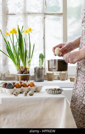 Preparazione per la Pasqua. Mani femminili con colore uovo di pasqua sotto il piatto vicino a tovaglia bianca decorata tabella marrone uova colorate e fiori di colore giallo con wi Foto Stock