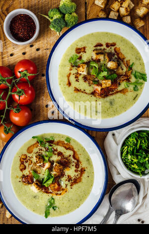 La crema di broccoli minestra in due ciotole bianco, guarnita con una polvere di peperoncino Salsa, crostini e prezzemolo. Accompagnata da cucchiai, extra di prezzemolo tritato, re Foto Stock