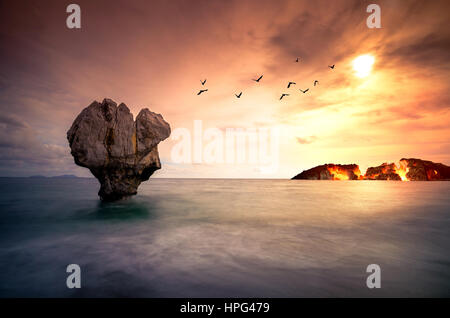 Arte con lonely scultura in pietra nel mare con sagome di uccelli in volo e un'isola che brucia sotto il tramonto. Foto Stock