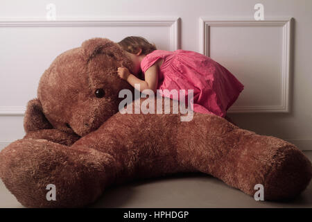 Orizzontale vista anteriore di un po' di Caucasian ragazza con i capelli corti e un abito rosa sussurra all'orecchio di un marrone grande orso di peluche su un pavimento contro Foto Stock