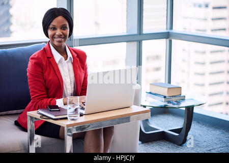Femmina nero imprenditrice sorridente con la videocamera mentre si è seduti su un lettino in una business lounge con le sue mani sul suo computer portatile e la tastiera con grandi finestre Foto Stock