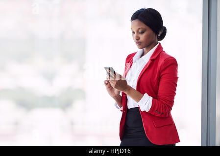 Giovani formalmente vestiti donna adulta occupata utilizzando il suo telefono cellulare mentre in piedi di fronte a grandi finestre di vetro mentre si indossa un rosso brillante blazer con c Foto Stock