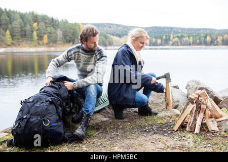 Giovane uomo con zaino guardando alla donna il taglio di legno sul lungolago durante il campeggio Foto Stock