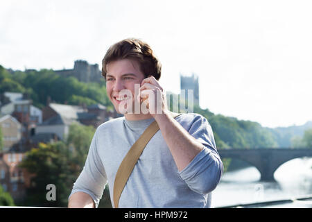 Un giovane uomo utilizza il suo telefono cellulare all'aperto vicino a un fiume. Egli sta parlando con qualcuno. Una con la cattedrale possono essere visti sullo sfondo. Foto Stock