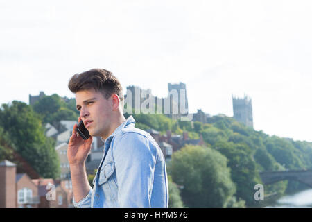 Un giovane uomo utilizza il suo telefono cellulare all'aperto vicino a un fiume. Egli sta parlando con qualcuno. Una con la cattedrale possono essere visti sullo sfondo. Foto Stock