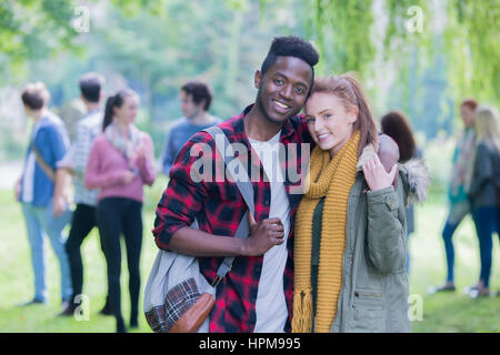 Una felice coppia giovane sorride come rilassarsi all'aperto con i loro amici. Essi sono su una pausa dallo studio in piedi in un abbraccio. Foto Stock