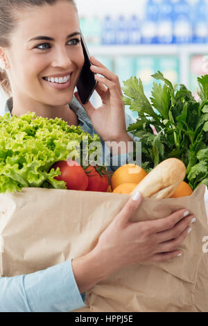 Bella giovane donna fare la spesa al supermercato, sta avendo una telefonata e tenendo un sacchetto con verdure fresche Foto Stock