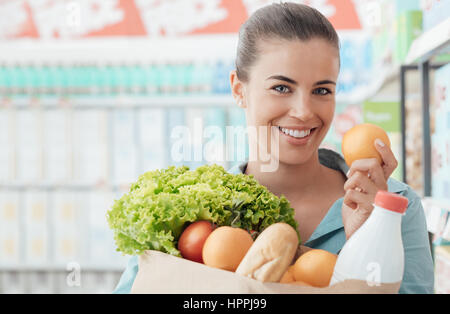 Sorridente giovane donna fare la spesa al supermercato, lei sta tenendo in mano un sacchetto con verdure fresche, pane e latte Foto Stock