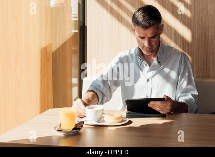 Fiducioso imprenditore avente la colazione presso la caffetteria e il lavoro con un touch screen tablet Foto Stock