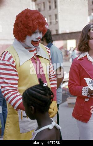 Una ragazza afro-americana passeggiate passato un attore vestito come il personaggio Ronald McDonald nel Bronx, New York, New York, 1976. Foto Stock