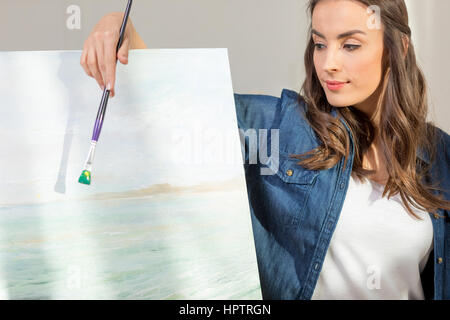 Bella giovane donna artista con il pennello appoggiata al cavalletto con foto Foto Stock