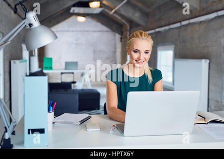 Giovane donna sorridente e digitando su laptop in un ufficio moderno Foto Stock