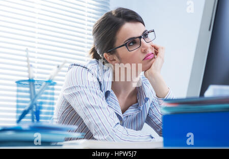 Annoiato lavoratore di ufficio alla scrivania a fissare lo schermo del computer con una mano sul mento. Foto Stock