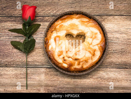 Torta di mele con un incavo a forma di cuore su di un tavolo di legno in cui vi è una rosa rossa Foto Stock
