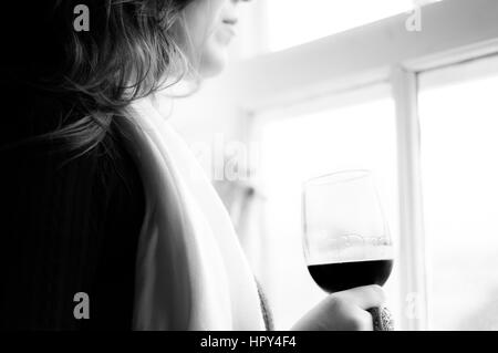 Un elegante donna in piedi da una finestra con in mano un bicchiere di vino rosso. Foto Stock