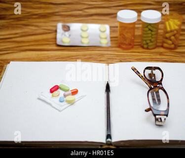 Un vecchio libro, coppia di rottura vetri lettore sullo sbozzato ufficiale e penna, sul tavolo di legno, circondato da un mucchio di pillole, suggerendo una persona anziana wit Foto Stock
