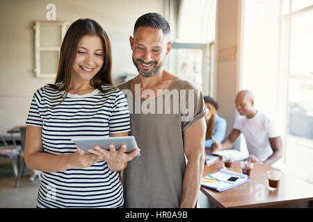 Giovane donna in piedi e mostrando tablet all uomo ed entrambi sorridente in camera con co-lavoratori. Foto Stock