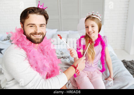 Mangiare e sorriso. Bel uomo barbuto guardando dritto sulla fotocamera indossando divertenti accessori rosa azienda rosso caramella in forma di stella nella mano destra Foto Stock