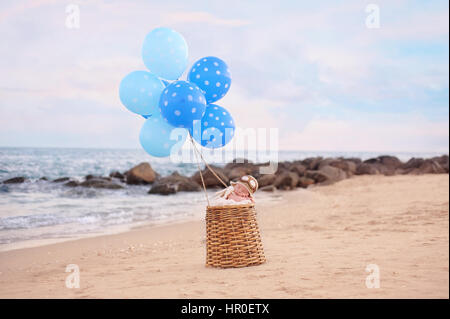 Due settimane vecchio neonato Bambino addormentato in un cesto di vimini con palloncini blu. Indossa un aviatore hat. Fotografato su una spiaggia con una roccia jetty. Foto Stock