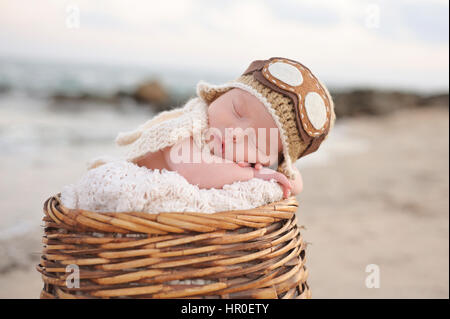 Due settimane vecchio neonato Bambino addormentato in un cesto di vimini indossando un aviatore hat. Fotografato su una spiaggia con una roccia jetty. Foto Stock