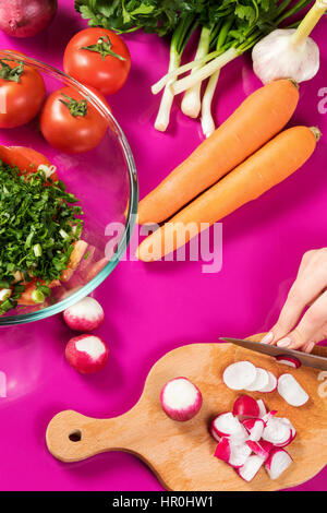 Mani femminili ravanelli tagliati per insalata su una tabella di colore rosa piena di verdure Foto Stock